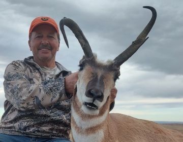 A hunter in a camouflage jacket and orange hat holds a large pronghorn antelope with unusal horns, celebrating a memorable hunt with SNS Outfitter & Guides under a moody sky.